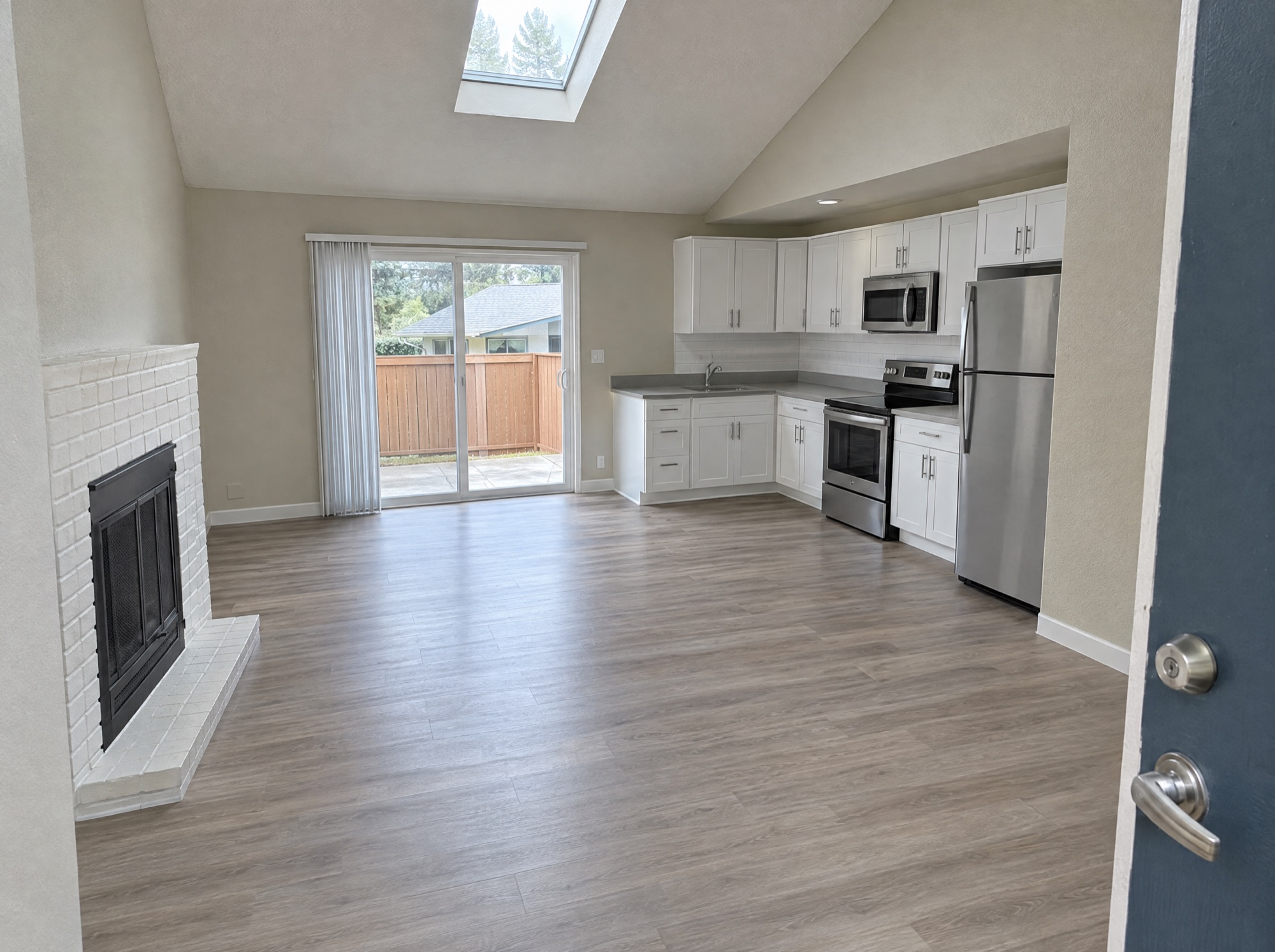 Vacant apartment seen from the entry door: vaulted ceiling with a skylight, sliding glass door with vertical blinds, white kitchen cabinets and stainless appliances